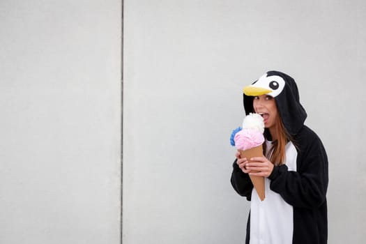 beautiful, young woman in penguin costume is eating decoration ice cream in front of concrete wall