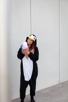 beautiful, young woman in penguin costume is eating decoration ice cream in front of concrete wall