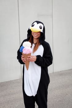 beautiful, young woman in penguin costume is eating decoration ice cream in front of concrete wall