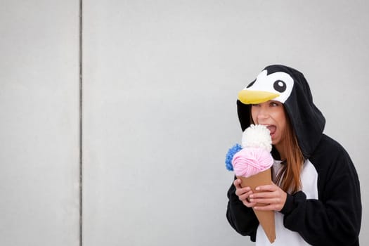 beautiful, young woman in penguin costume is eating decoration ice cream in front of concrete wall