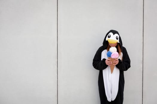 beautiful, young woman in penguin costume is eating decoration ice cream in front of concrete wall