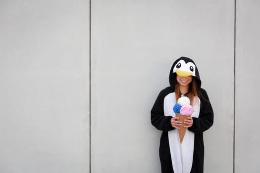 beautiful, young woman in penguin costume is eating decoration ice cream in front of concrete wall