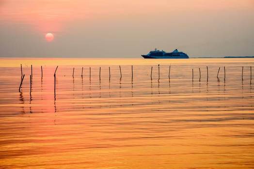 Travel by Cruise ship in the ocean, large luxury passenger boat is sailing on the bright sea, red sun in the colorful sky is yellow, orange, beautiful nature landscape at sunset or sunrise background
