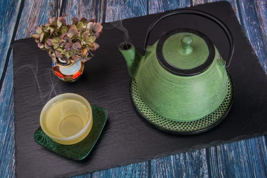 A steaming green metal teapot, a glass bowl with steaming tea and a chinese vase with dried hydrangea on a slate slab.