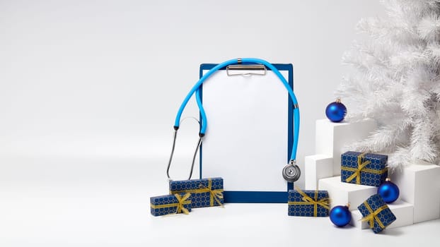 Stethoscope, blank clipboard, white fir tree, gift boxes and Christmas decorations on white background with copy space. Medical concept. Greeting card. New Year and Christmas.