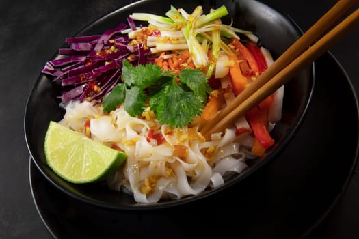 Spicy rice noodel salad in a black bowl with chopsticks, including peppers, cabbage, scallions, and chili lime dressing