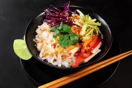 Spicy rice noodel salad in a black bowl with chopsticks, including peppers, cabbage, scallions, and chili lime dressing