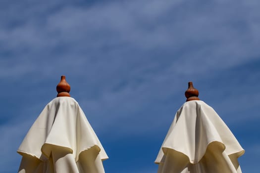 Sun umbrellas beach. Close-up of two closed white beach umbrellas or parasols against blue cloudy sky with selective focus. Macro photograph. Tourism and travel destination concept. Space.