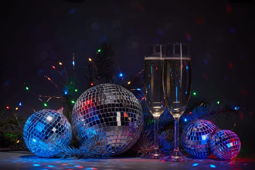 Champagne flutes, mirror balls (disco ball), fir branch, on dark background with colorful LED lights garland. New Year and Christmas. Weekend or holiday party.