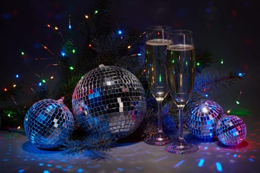 Champagne flutes, mirror balls (disco ball), fir branch, on dark background with colorful LED lights garland. New Year and Christmas. Weekend or holiday party.
