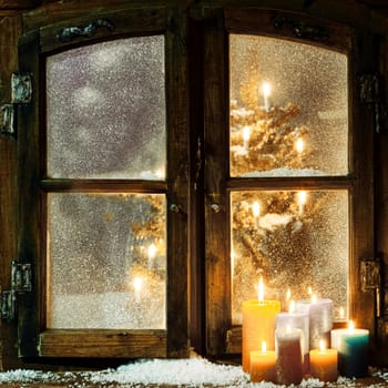 Welcoming Christmas window in a log cabin with a group of burning candles on the windowsill and a glowing Christmas tree visible through the frosted panes