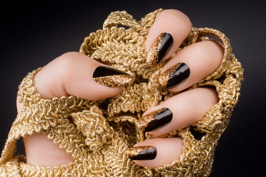 Female hand with golden black nails is holding a golden decoration on black background.