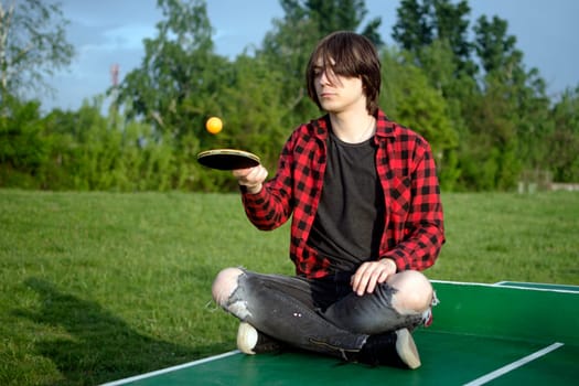 The teen boy is sitting on the green table for table tennis and playing alone with racket and ball outdoors.