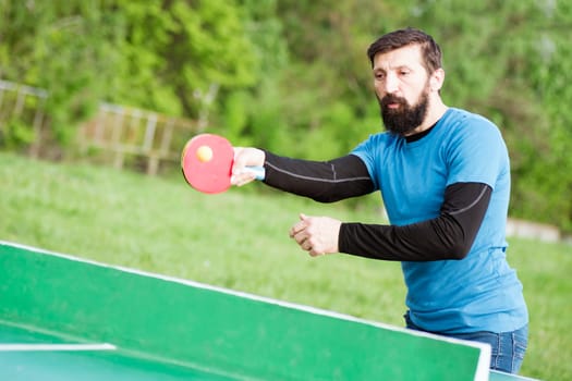 The bearded player is playing table tennis as recreation outdoors in the park.