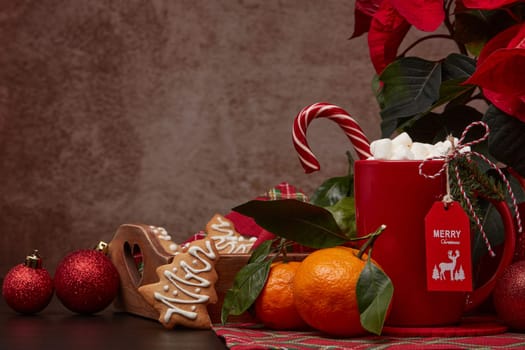 Red Poinsettia flowers, tray with ginger cookies and mandarines, mug with hot cocoa and marshmallows. fir ornaments on brown background with copy space. Winter holidays. New Year, Christmas treat.