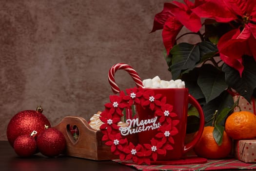 Red Poinsettia flowers, tray with ginger cookies, mandarines, mug with hot cocoa and marshmallows. fir ornaments, gifts on brown background with copy space. Winter holidays. New Year, Christmas treat