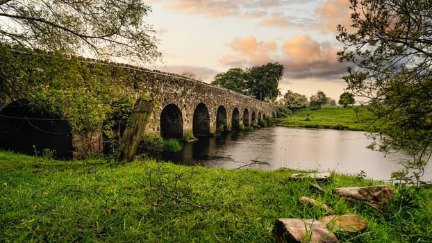 Old 12th century stone arch bridge over a river, rocks on first plane. Green fields and trees. Dramatic sky sunset. Count Meath, Ireland