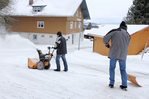 Two men clear the road and the sidewalk with a snow blower and snow shovel