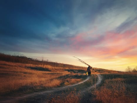 Old man silhouette walking a country road carrying dry tree branches on his shoulders, against sunset sky background. Idyllic rural landscape, late autumn season and dirt track across the golden hay.