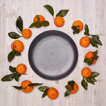 Frame from ripe mandarins with leaves and black empty plate on light wooden background. Top view with copy space. Festive background from tangerines