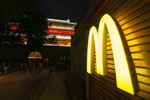 Xian, China -  July 2019 : Yellow lit Macdonald restaurant logo at night, with landmark Drum Tower heritage building in the background