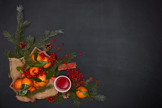 Festive still life: berries, mandarins with leaves on wooden tray, fruit tea on red mug, spices and fir branches on dark background. Top view with copy space. Christmas. New Year