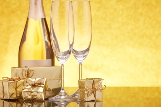 Golden gift boxes, champagne bottle and empty champagne flutes on festive gold background. Christmas. New Year. Valentine day. Wedding. Design element for greeting card, invitation, advertising