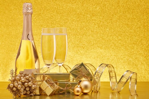 Golden Christmas ornaments, gift boxes, champagne bottle and full champagne flutes on gold background with copy space. Christmas. New Year. Design element for greeting card, invitation, advertising