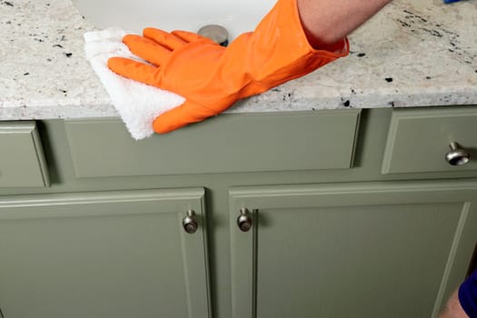 Granite bathroom counter being cleaned with a white cotton cleaning rag by a man wearing an orange rubber glove. Stone counter being cleaned with a rag by a man wearing an orange protective glove.