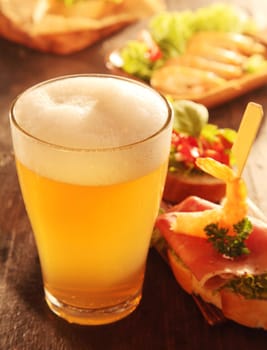 Buffet of assorted gourmet tapas with shrimp, ham and tomato on a wooden table with a glass of cold frothy beer, close up high angle view with focus to the froth