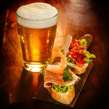 Glass of cold frothy beer with gourmet appetizers consisting of prawns, prosciutto ham and tomato on sliced baguette served on a wooden bar counter
