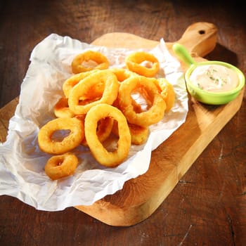 Crispy golden deep fried squid or calamari rings in batter served with a savory dipping sauce on a wooden board, closeup high angle view