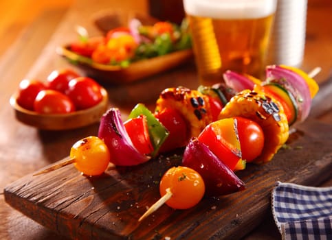Colorful vegetarian or vegan kebabs with fresh veggies including sweet bell peppers, mushroom, carn on the cob, onion and tomato served with a side salad and beer on a wooden board in a pub