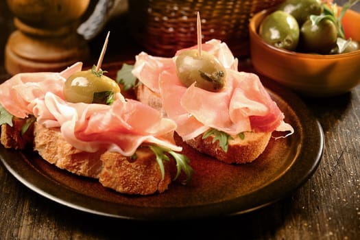 Canapes or tapas with proscuitto ham and olives on crusty fresh baguette served as appetizers or snacks at a party