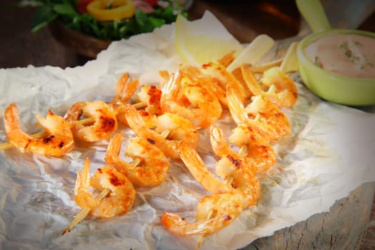 Three gourmet appetizers of crispy grilled prawns in garlic on skewers served on crumpled white paper with a lemon garnish and savory dip