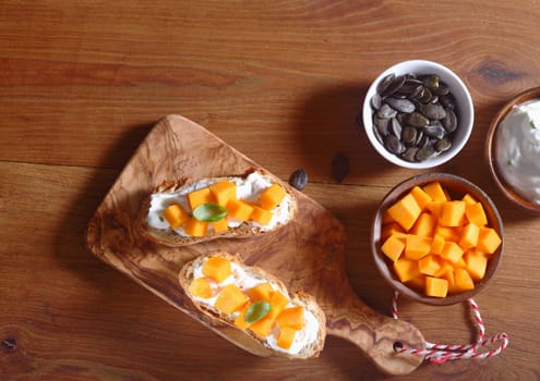 Close up Aerial Shot of Delicious Crusty Bread with Cream and Carrot Slice Fillings on Top of Wooden Board. Placed on Wooden Table Bowl of Carrot Slices and Pumpkin Seeds on the Side.