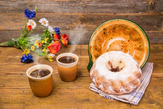 A Gugelhupf with its ceramic baking dish and two cups of coffee and a field flowers birthday bouquet on a wooden table with a wooden background.
