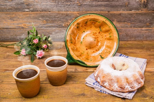 A Gugelhupf with its ceramic baking dish and two cups of coffee and a field flowers birthday bouquet on a wooden table with a wooden background.