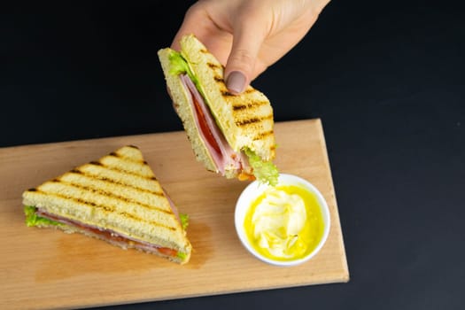 Sandwich of several slices of white bread on a wooden board. The filling of the sandwich consists of bacon, cheese and ham. Next cutlery and a cup of mustard. Dark wooden background. View from above.