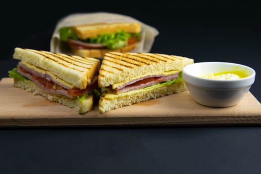 Sandwich of several slices of white bread on a wooden board. The filling of the sandwich consists of bacon, cheese and ham. Next cutlery and a cup of mustard. Dark wooden background. View from above.