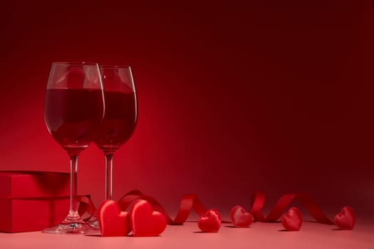 Gift box, red hearts, two glasses with wine on red background. Valentine's Day card. Copy space. Design element for romantic greeting card, wedding invitation.