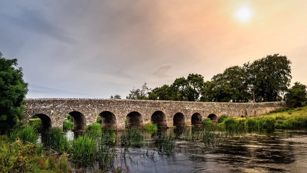 Old 12th century stone arch bridge over a river. Green fields and trees. Dramatic sky at sunrise. Count Meath, Ireland