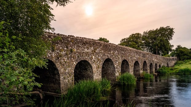 Old 12th century stone arch bridge over a river. Green fields and trees. Dramatic sky at sunrise. Count Meath, Ireland