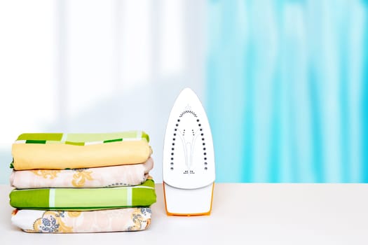 Household laundry ironing. Close-up of a yellow electrical iron and a stack of ironed clothes on white board over blurred curtains background. Advertising laundry service.