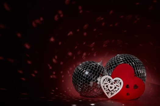 Glittering mirror disco ball  and decorative hearts on dark background with red lighting. Copy space. Valentine's Day background. Holiday party. Nightclub. Entertainment, disco, music show background