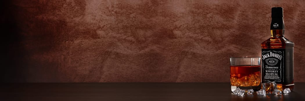 St.Petersburg, Russia - December 2019 - Bottle of Jack Daniel's whiskey and glass with drink and ice on wooden table on brown background with copy space. Extra wide panorama banner background