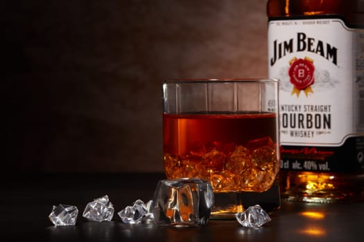St.Petersburg, Russia - December 2019 - Bottle of Jim Beam bourbon whiskey and glass with drink and ice  on wooden table on brown background with copy space. Kentucky straight bourbon whiskey
