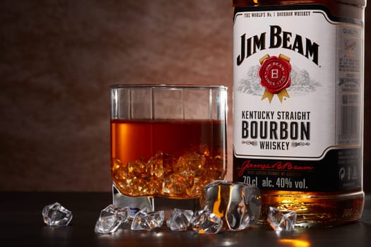 St.Petersburg, Russia - December 2019 - Bottle of Jim Beam bourbon whiskey and glass with drink and ice  on wooden table on brown background. Kentucky straight bourbon whiskey