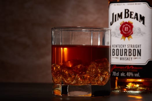 St.Petersburg, Russia - December 2019 - Bottle of Jim Beam bourbon whiskey and glass with drink and ice  on wooden table on brown background. Kentucky straight bourbon whiskey
