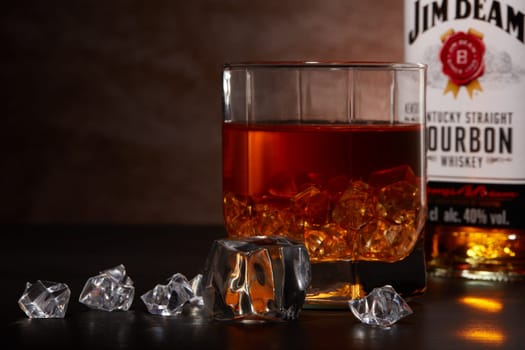 St.Petersburg, Russia - December 2019 - Bottle of Jim Beam bourbon whiskey and glass with drink and ice  on wooden table on brown background. Kentucky straight bourbon whiskey